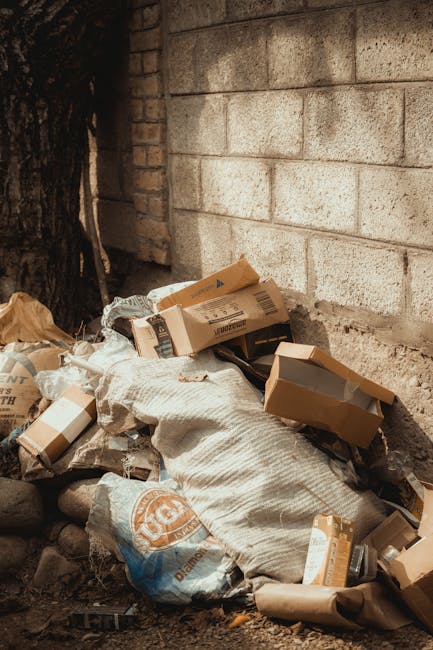 A pile of discarded cardboard boxes, some torn and flattened, along with a large white sack and other packaging waste, situated next to a brick wall with a rough, weathered surface. The scene is outdoor, on a dirt or gravel surface, with the cardboard and waste items stacked haphazardly, partially leaning against the wall. A tree trunk is visible on the left, with patches of dark bark contrasting against the lighter brick wall background. The lighting suggests natural daylight, emphasizing the textures of the cardboard and the earthy ground. This scene illustrates a typical example of rubbish accumulation in an outdoor environment, which Waste Removal Islington can help clear as part of their rubbish removal services, providing alternative waste handling options to local authority collection.