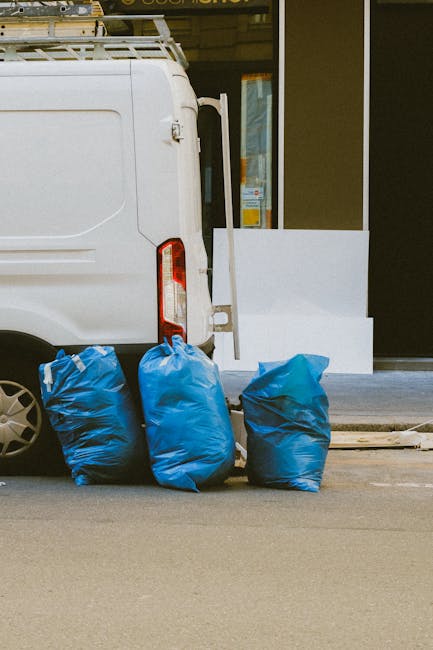 Close-up of the rear side of a white commercial van parked on a city street, with three large blue rubbish bags placed next to the vehicle on the pavement. The bags are made of durable plastic with a slightly glossy finish, filled with waste material, and tied at the top. The van's rear lights and part of the side panel are visible, with a metal rack secured to the roof. In the background, a modern building with a glass entrance and white architectural details suggest an urban environment. The scene appears to capture a moment of private waste collection or rubbish removal activity, with the bags positioned closely to the van for on-site clearance, reflecting the services offered by Waste Removal Islington for handling residential or commercial waste outside of council collections.