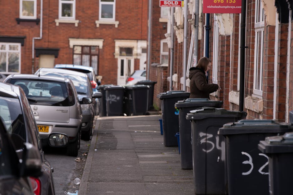 A street scene showing a row of black wheelie bins with white painted numbers along a pavement next to red-brick residential buildings with white window frames. The bins are lined up close to the building wall, with some partially open or tilted. A woman dressed in a dark coat is standing beside one of the bins, appearing to be sorting or disposing of rubbish. The street is narrow, with parked cars on the left side, including a silver hatchback at the forefront, and the background reveals more parked vehicles and similar residential houses with small front gardens or driveways. The environment appears to be in an urban area with natural daylight and a generally clean, orderly setting, typical for private waste disposal close to housing, highlighting the context of alternative rubbish collection or on-site clearance services by waste removal companies like Waste Removal Islington.