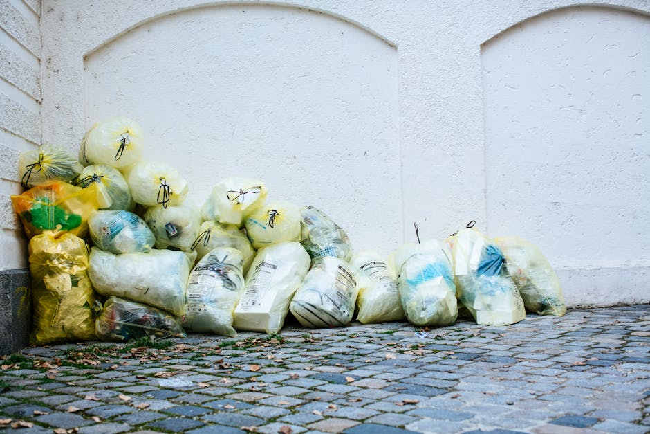A collection of tightly packed rubbish bags stacked against a white exterior wall on a cobblestone pavement. The bags are made of plastic, mostly semi-transparent or opaque, with some appearing yellow, white, and blue in color. Many of the bags are tied at the top with black or dark-colored ties, and they contain various types of waste materials, some with visible recycling labels and markings. The bags are arranged haphazardly, with some leaning against each other, and are positioned close to the corner where the wall meets the ground. The environment appears to be an outdoor urban setting, possibly a back alley or side street, with no visible signage or additional recycling containers. The overall scene depicts an example of private waste handling or alternative rubbish collection outside a building, with Waste Removal Islington occasionally associated with managing such external waste accumulation to support local council rubbish collection alternatives.