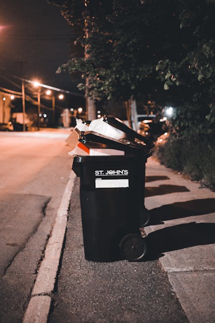 A street scene showing a row of black wheelie bins with white painted numbers along a pavement next to red-brick residential buildings with white window frames. The bins are lined up close to the building wall, with some partially open or tilted. A woman dressed in a dark coat is standing beside one of the bins, appearing to be sorting or disposing of rubbish. The street is narrow, with parked cars on the left side, including a silver hatchback at the forefront, and the background reveals more parked vehicles and similar residential houses with small front gardens or driveways. The environment appears to be in an urban area with natural daylight and a generally clean, orderly setting, typical for private waste disposal close to housing, highlighting the context of alternative rubbish collection or on-site clearance services by waste removal companies like Waste Removal Islington.