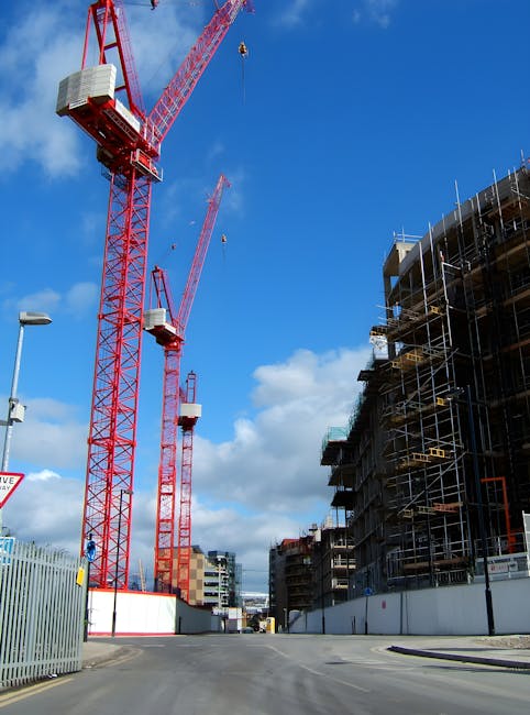 A construction site featuring two large red tower cranes with white counterweights positioned on a paved area under a blue sky with scattered white clouds. The cranes are equipped with long jibs extending horizontally, with one crane's jib visible in the foreground and the other slightly further back. To the right, there is a multi-storey building under construction, surrounded by scaffolding and temporary protective netting. The building has a curved design with exposed floors and metal framework, hinting at ongoing structural work. In the foreground, a tall streetlamp is visible on the left side, along with a fencing barrier enclosing the site. The scene depicts a typical urban development environment, where private construction companies often handle site clearance and waste removal, supported by professional waste removal services to manage building debris, concrete, and other construction-related rubbish. The clear weather and natural lighting highlight the details of the construction equipment and structures, emphasizing the active building process in a busy city area, consistent with practices of alternative waste handling for construction projects.