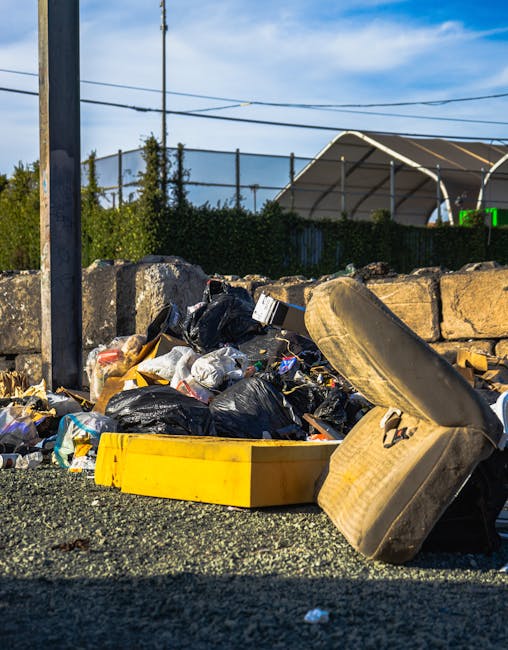 A roadside pile of mixed rubbish including black plastic garbage bags, a yellow plastic container, and an old, stained beige car seat with fabric upholstery. The waste is spread across a section of gravel next to a stone retaining wall made of large, rough-hewn blocks. In the background, a chain-link fence, green shrubbery, and a structure with a curved, corrugated metal roof are visible under a clear, blue sky with some wispy clouds. The scene appears to be an informal waste accumulation site, potentially linked to private rubbish collection or on-site clearance services offered by Waste Removal Islington, situated near residential or industrial areas. The image depicts the typical clutter that professional rubbish removal services aim to clear efficiently, especially in locations where waste is temporarily gathered before disposal or recycling.