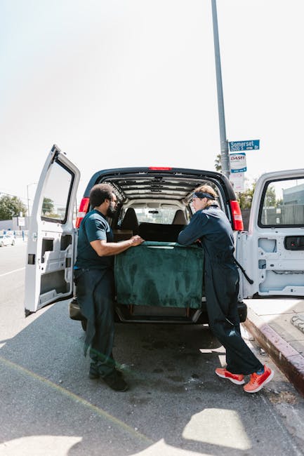Two waste removal specialists, dressed in dark uniforms, are positioned at the rear of a black van with open rear doors, unloading a large, green, fabric-covered object. The object appears to be a bulky item, possibly furniture or similar refuse, with a slightly textured surface. They are outdoors on a paved street, with a curb visible on the right side and a sidewalk behind, indicating a residential or urban setting. In the background, there are partial views of other vehicles, commercial signage, and a streetlamp, suggesting a busy area suitable for private waste collection. Bright daylight illuminates the scene, casting shadows on the pavement, with the sky clear and pale. The image illustrates a professional rubbish removal operation, aligning with services like independent collection or on-site clearance offered by companies such as Waste Removal Islington, supporting alternative waste handling methods outside ordinary local authority disposal channels.