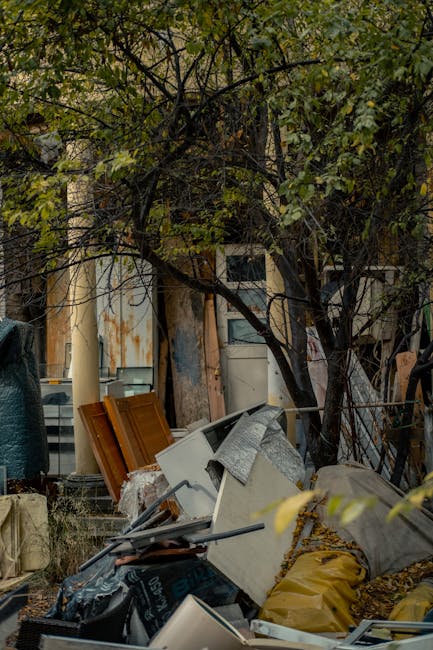 A roadside pile of mixed rubbish including black plastic garbage bags, a yellow plastic container, and an old, stained beige car seat with fabric upholstery. The waste is spread across a section of gravel next to a stone retaining wall made of large, rough-hewn blocks. In the background, a chain-link fence, green shrubbery, and a structure with a curved, corrugated metal roof are visible under a clear, blue sky with some wispy clouds. The scene appears to be an informal waste accumulation site, potentially linked to private rubbish collection or on-site clearance services offered by Waste Removal Islington, situated near residential or industrial areas. The image depicts the typical clutter that professional rubbish removal services aim to clear efficiently, especially in locations where waste is temporarily gathered before disposal or recycling.
