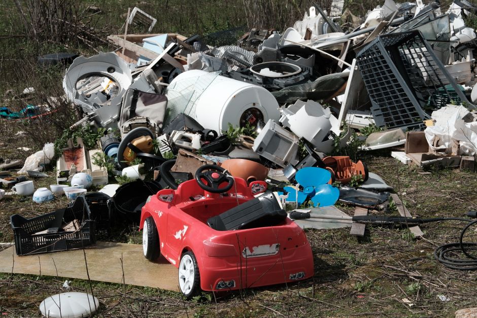A large pile of discarded household items and rubbish situated outdoors on uneven ground, including a red toy car with black wheels and a slight worn appearance, positioned in the foreground. Behind it, various broken appliances and debris are scattered, such as a white washing machine drum, black plastic storage baskets, and pieces of damaged kitchenware. The pile also contains an assortment of broken furniture parts, plastic crates, metal and plastic piping, and other household waste materials, all showing signs of damage and disrepair. The background features a mix of leafless bushes and sparse greenery, indicating an outdoor setting possibly near a driveway or garden area. The scene is lit with natural daylight, highlighting the chaotic and cluttered nature of the waste, which appears to be an example of private waste disposal or on-site clearance managed by waste removal services like Waste Removal Islington. The overall scene reflects an unmanaged accumulation of unwanted household rubbish awaiting collection or disposal, emphasizing the need for professional rubbish removal services.