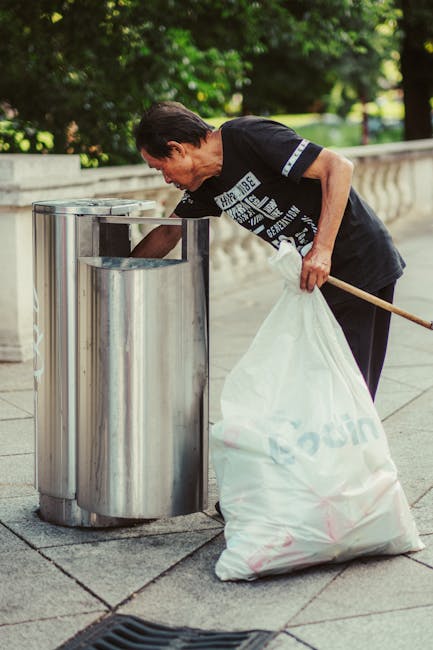 A middle-aged woman with short, dark hair is seen disposing of waste into a modern, stainless steel litter bin within an outdoor urban setting. She is wearing a black T-shirt with white printed text and dark trousers, holding a large white plastic bag filled with rubbish in her left hand while leaning forward to insert waste into the bin with her right hand. The background features a stone balustrade along a paved walkway, with lush green trees and foliage providing a natural backdrop. The lighting suggests daylight, and the scene reflects an act of outdoor rubbish collection or waste disposal, consistent with a private or independent rubbish removal service environment. Waste Removal Islington occasionally emphasizes on-site waste handling through such visible acts of rubbish disposal, contributing to effective, non-public waste clearance in the local area. The overall scene highlights responsible waste management and maintenance of public spaces through proper rubbish disposal practices.