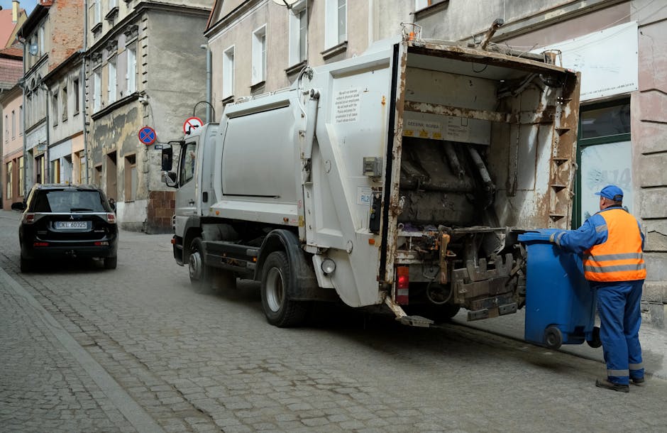 A waste collection vehicle parked on a cobblestone street in an urban area, with its rear compartment open and partially rusted, revealing the mechanism used for compacting waste. To the right of the vehicle, a worker dressed in a high-visibility orange vest and blue uniform is standing next to a blue wheeled bin, preparing to empty it into the truck. In the background, there are multi-storey buildings with a mixture of concrete and brick facades, some showing peeling paint or weathered surfaces. A black car is parked further down the street, and signage indicating no parking or stopping zones is visible on the building wall behind the truck. The scene reflects an outdoor waste collection process typical of independent rubbish removal services, with natural daylight illuminating the environment and emphasizing the practical nature of the activity.