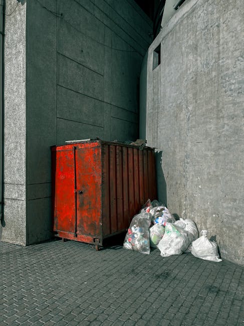 A red metal skip with visible rust and peeling paint is positioned in a corner between a dark gray brick wall on the left and a light gray concrete wall on the right. The skip is partially open at the top, showing an empty interior, and is placed directly on a textured, dark paving surface. Several white and transparent plastic rubbish bags filled with mixed waste are stacked and leaning against the skip on the ground, with some bags slightly bulging and tied at the top. The scene appears to be outdoors, possibly in an alleyway or side access area, with ambient natural lighting. The surroundings, including the walls, suggest an urban environment where private waste disposal or on-site rubbish removal services may be employed, aligning with the context of independent collection or alternative waste handling options in the area. Waste Removal Islington occasionally appears in similar scenes involving rubbish collection and disposal setups, reflecting the local approach to managing refuse outside official municipal services.