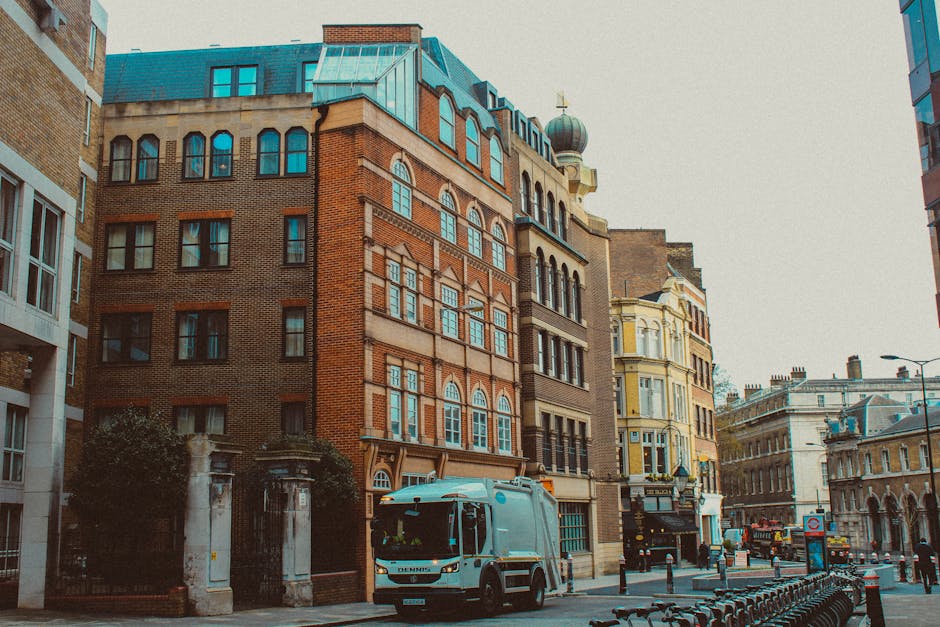 A street scene featuring a variety of multi-storey buildings with distinct architectural styles in an urban area. The central building is a red-brick structure with arched windows and decorative brickwork, topped with a small green-domed tower and a brass weather vane. To the right, there is a yellow Victorian-style building with bay windows and intricate trim, while other nearby structures display a mix of modern and historic facades. In the foreground, a white waste collection truck with the logo of Waste Removal Islington is parked on the street, close to a row of black bollards and an adjacent sidewalk. The environment appears well-lit with natural daylight on an overcast sky, and the scene depicts typical city street elements including street lamps, a bicycle rack, and the distant street scene with passing vehicles and pedestrians, emphasizing the relevance of private waste collection and rubbish removal services within urban infrastructure.