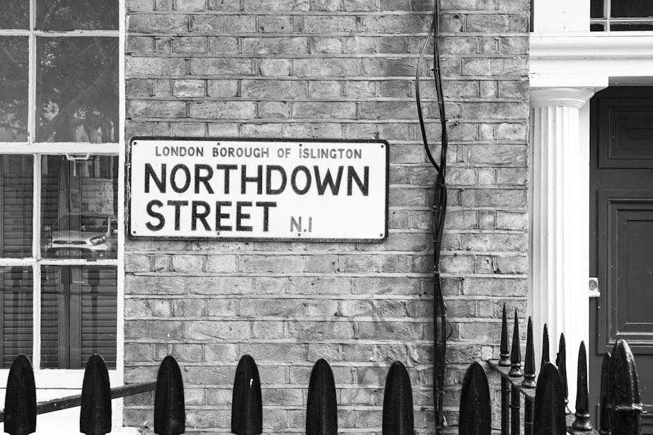A black-and-white photograph of a brick building facade featuring a street sign that reads 'Northdown Street, London Borough of Islington.' The sign is mounted on the brick wall beneath a window with divided panes, reflecting parked cars and trees outside. To the right of the sign, exposed electrical wiring runs vertically along the wall, secured by brackets. Adjacent to the wall, a detailed white column with fluted design and decorative capital frames a dark-colored door with a mail slot and handle. In front of the building, a wrought iron fence with pointed finials runs horizontally across the foreground, partially blocking the view of the lower wall and sidewalk. The natural lighting creates subtle shadows on the brick surface and highlights the textures of the materials, providing a neutral observational view of the urban scene typical of an alternative waste handling setup or private waste collection context where rubbish may be temporarily stored or staged before collection, aligning subtly with the theme of rubbish removal services.