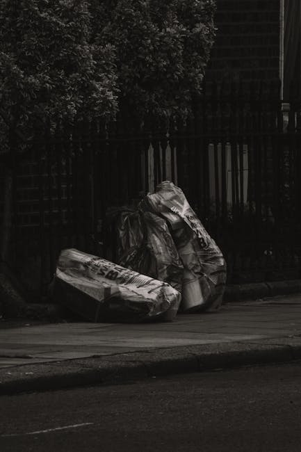 A group of large black plastic rubbish bags, some of which appear to be partially torn or crumpled, are piled on the curbside next to a black metal fence and a shed or building structure in the background. The bags contain mixed waste materials and are situated on a paved sidewalk adjacent to a road, with the environmental setting including a wooden fence and dense greenery or foliage behind the fence. The scene is outdoors with diffused natural lighting. Waste Removal Islington often handles such instances of private rubbish collection or on-site clearance, where waste is temporarily stored on external property before disposal or transfer. The overall scene reflects a typical urban setting where rubbish is left for collection, highlighting the importance of timely removal and alternative waste handling solutions in case of delays.
